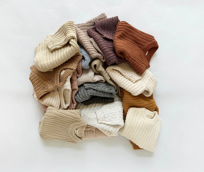 May include: A collection of folded long-sleeve sweaters in various neutral earth tones including cream, beige, tan, brown, grey, and dark purple. Each sweater features a thick, chunky knit waffle texture with a crew neckline and ribbed cuffs.