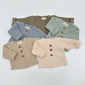May include: Five knitted baby cardigans in different colors: light brown, blue, green, beige, and a darker brown. Each cardigan has a button closure and a simple design.