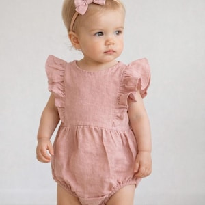 May include: A baby wearing a dusty rose-colored linen romper with ruffled sleeves and a matching bow headband. The romper has a bubble silhouette. The baby is standing and looking to the side.