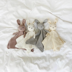 May include: Four crocheted bunny security blankets in shades of mauve, white, gray, and cream. Each bunny has closed eyes and an embroidered 'X' for a mouth. The blankets are lying on a white sheet.