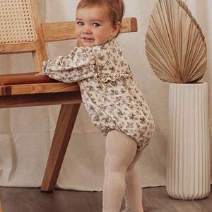May include: A baby wearing a floral print long-sleeved onesie and beige tights. The baby is standing next to a wooden chair and a tall vase with a dried palm leaf.