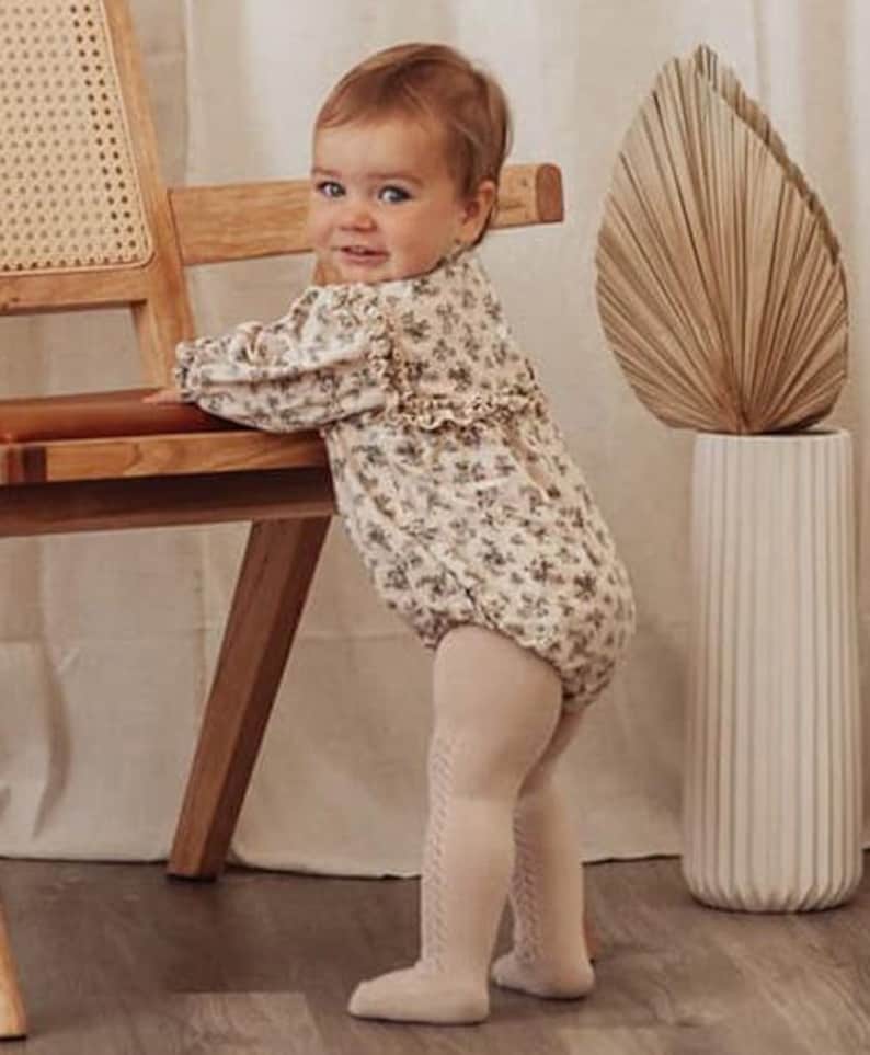 May include: A baby wearing a floral print long-sleeved onesie and beige tights. The baby is standing next to a wooden chair and a tall vase with a dried palm leaf.