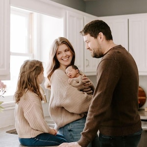 May include: A family of four in a kitchen. A child with long hair wears a beige knit sweater and blue jeans. A parent with long hair wears a beige knit sweater and blue jeans, holding a newborn in a beige knit outfit. A parent wears a brown sweater.