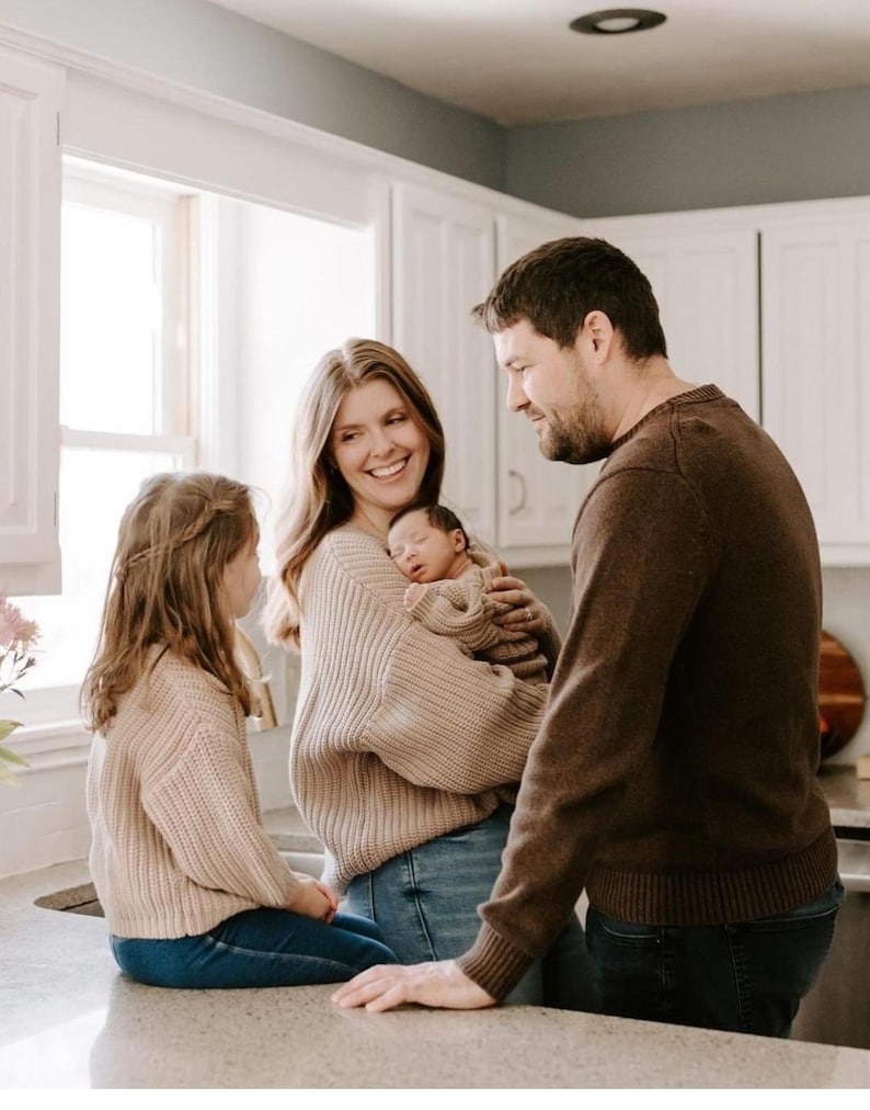 May include: A family of four in a kitchen. A child with long hair wears a beige knit sweater and blue jeans. A parent with long hair wears a beige knit sweater and blue jeans, holding a newborn in a beige knit outfit. A parent wears a brown sweater.