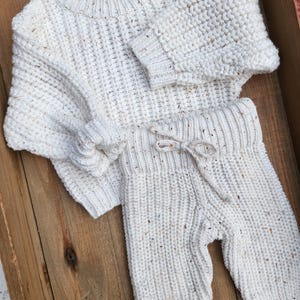 May include: A white knitted sweater and matching pants set with a speckled texture. The sweater has a crew neck and long sleeves. The pants have a drawstring waistband and a relaxed fit.