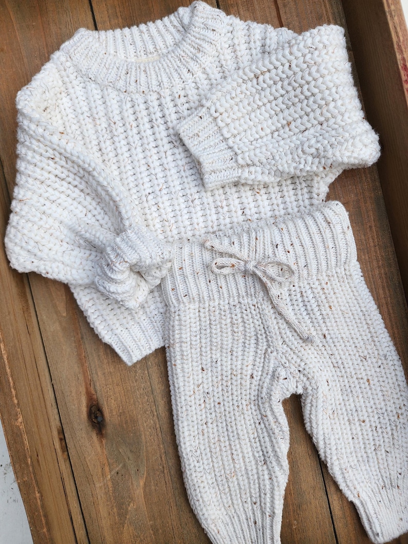 May include: A white knitted sweater and matching pants set with a speckled texture. The sweater has a crew neck and long sleeves. The pants have a drawstring waistband and a relaxed fit.