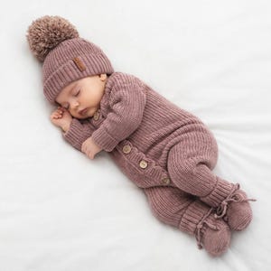 May include: A sleeping baby wearing a mauve knit romper with wooden buttons and matching hat with a pom-pom. The outfit includes attached booties with tie closures. The baby is lying on a white surface.