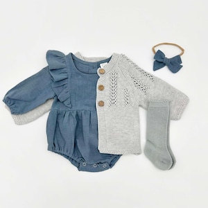 May include: A baby clothing set in shades of blue and gray, including a romper with ruffle details, a knit cardigan, socks, and a bow headband. The romper is a dusty blue, and the cardigan is light gray with wooden buttons.