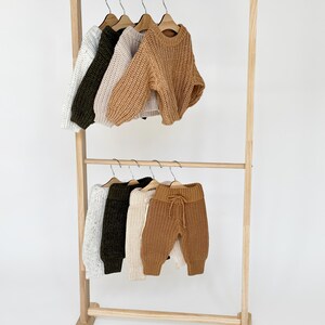 May include: A wooden clothing rack with two tiers, displaying a variety of knitted sweaters and pants in beige, white, and green.