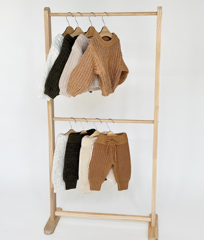 May include: A wooden clothing rack with two tiers, displaying a variety of knitted sweaters and pants in beige, white, and green.