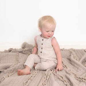 Op de afbeelding: Een peuter draagt een lichtbeige linnen jumpsuit met bruine knopen. De jumpsuit is mouwloos en heeft een ontspannen pasvorm. Het kind zit op een beige, getextureerd deken met franjes.