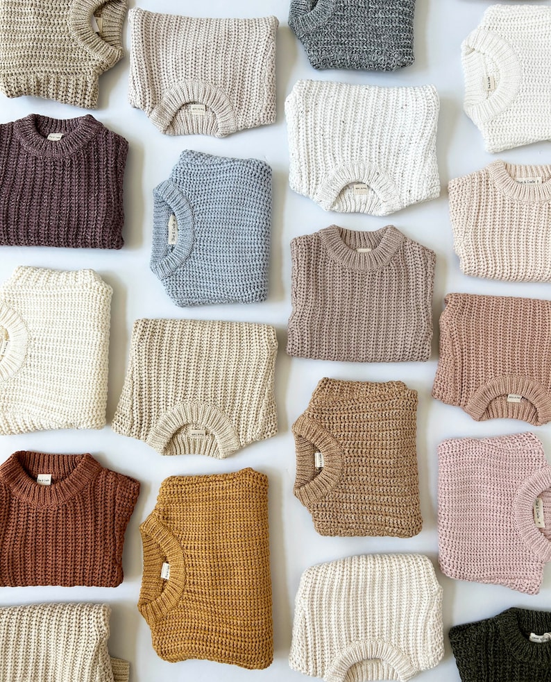 May include: A collection of folded long-sleeve sweaters in various solid colors including beige, brown, blue, white, gray, pink, and mustard. Each sweater features a thick, chunky knit texture, a ribbed crew neckline, and a small white brand label.