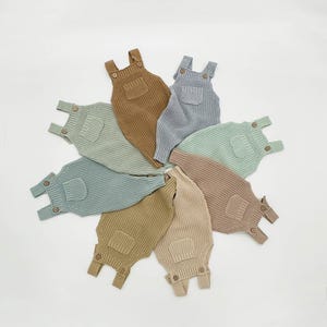 May include: Seven knitted baby dungarees in various colours, including light blue, brown, and beige. The dungarees have a pocket on the front and adjustable straps.