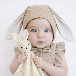 May include: A baby wearing a tan bunny ear bonnet and matching dress, holding a cream-colored crocheted bunny toy. The bunny toy has black stitched eyes and a pink blush. The baby has bright blue eyes.