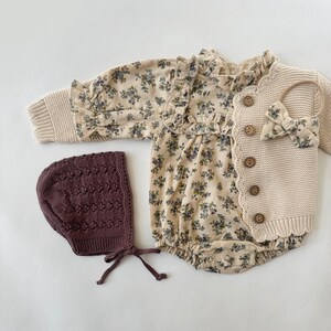 May include: A baby outfit set with a floral print romper, a knitted cardigan, and a brown knitted bonnet.