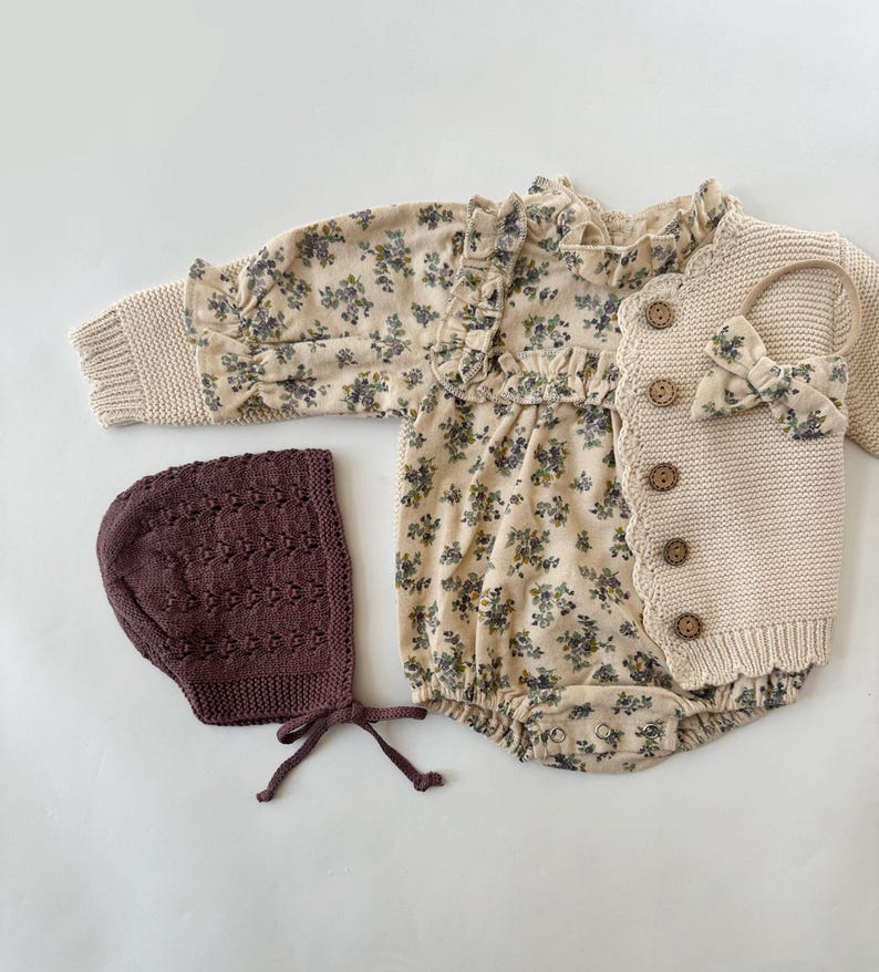 May include: A baby outfit set with a floral print romper, a knitted cardigan, and a brown knitted bonnet.