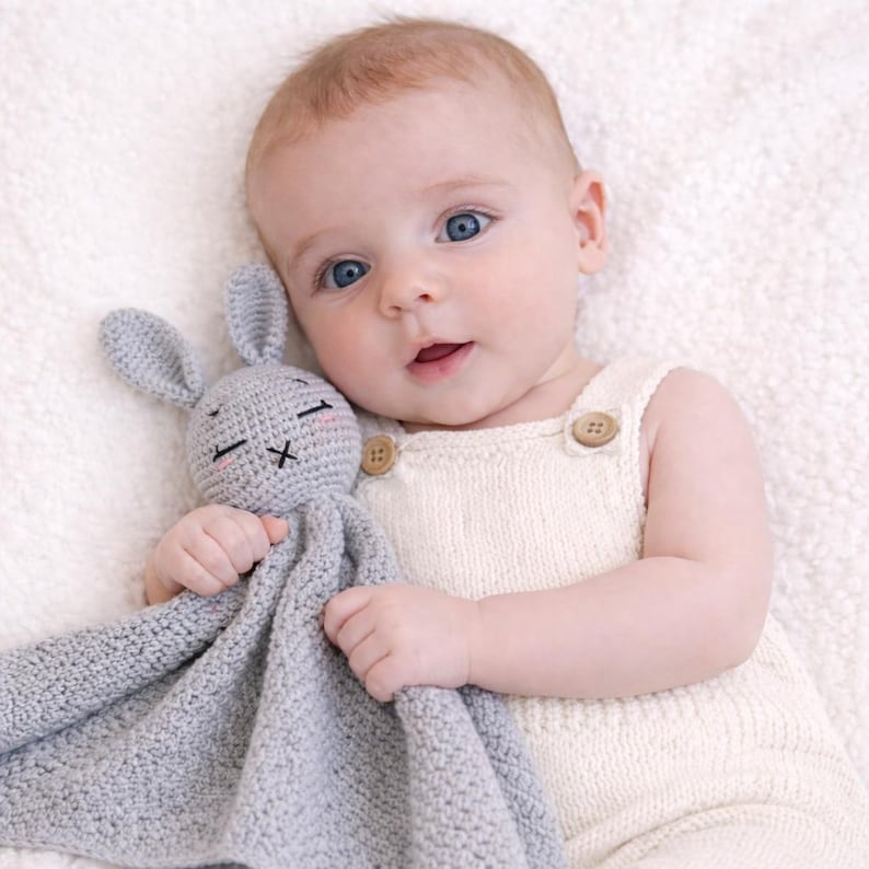 May include: A baby wearing a cream-colored knit romper with wooden buttons, holding a gray crocheted bunny lovey. The bunny has embroidered facial features and long ears. The baby has blue eyes and fair skin.