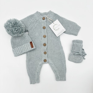 May include: A light blue knitted baby outfit set with a matching hat and booties. The outfit has a button closure and a pom-pom on the hat.