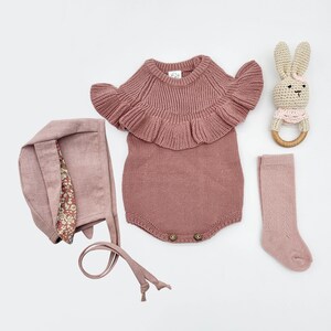 May include: A pink knitted baby onesie with a ruffled neckline, a pink knitted hat, a pair of pink socks, and a wooden bunny rattle.
