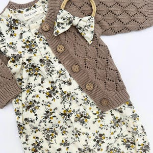 May include: A brown knitted cardigan with a diamond pattern and button fastening, a white floral romper, and a brown headband with a bow.