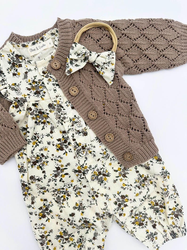 May include: A brown knitted cardigan with a diamond pattern and button fastening, a white floral romper, and a brown headband with a bow.