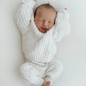 May include: A newborn infant is wearing a white, long-sleeved, chunky knit sweater with a matching pair of white knit pants. The sweater features a ribbed texture and a subtle speckled pattern throughout the fabric, with cuffed sleeves and a crew neckline.