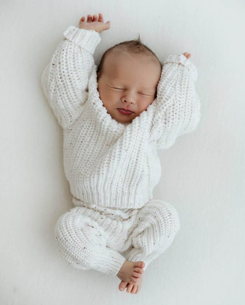 May include: A newborn infant is wearing a white, long-sleeved, chunky knit sweater with a matching pair of white knit pants. The sweater features a ribbed texture and a subtle speckled pattern throughout the fabric, with cuffed sleeves and a crew neckline.