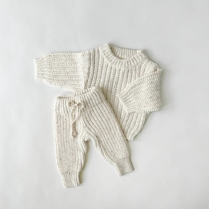 May include: A cream-colored knitted sweater and pants set for a baby or toddler. The sweater has a round neckline and long sleeves. The pants have an elastic waistband and a drawstring.
