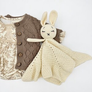 May include: A cream-colored crochet bunny lovey with closed eyes and a cross-stitched mouth rests beside a brown knit cardigan and a floral print onesie. The cardigan has wooden buttons and a diamond pattern.
