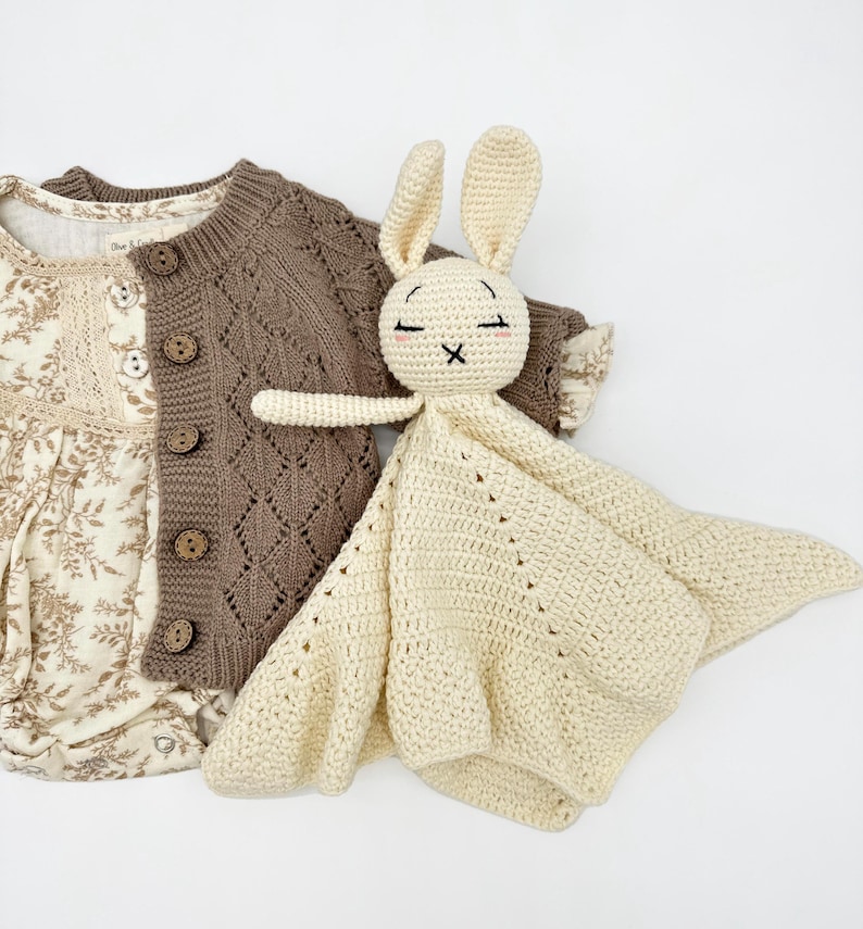 May include: A cream-colored crochet bunny lovey with closed eyes and a cross-stitched mouth rests beside a brown knit cardigan and a floral print onesie. The cardigan has wooden buttons and a diamond pattern.