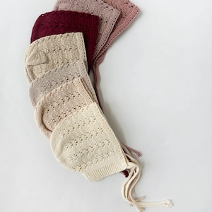 May include: A set of six crocheted baby bonnets in various shades of pink, beige, and burgundy. The bonnets have a delicate openwork pattern and are tied with long strings.