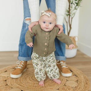 May include: A baby wearing a green cardigan with floral print trousers and a floral headband. The baby is standing on a woven rug.