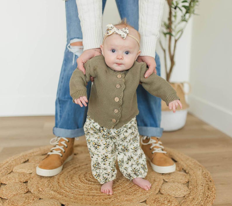 May include: A baby wearing a green cardigan with floral print trousers and a floral headband. The baby is standing on a woven rug.