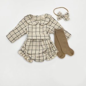 May include: A beige and black plaid baby outfit set, including a long-sleeved romper with a Peter Pan collar, a matching headband with a bow, and a pair of knee-high socks. The outfit is made of a soft fabric.