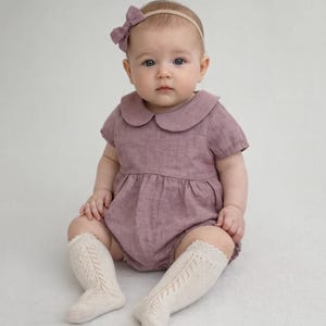 Puede incluir: Un bebé con un pelele de color malva con cuello Peter Pan y una diadema con lazo a juego. El bebé también lleva calcetines altos hasta la rodilla de color crema con un patrón calado decorativo.