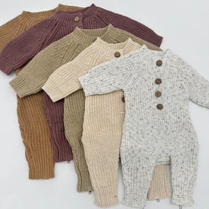 May include: Five knitted baby onesies in different colors, including beige, brown, cream, and white with speckles. The onesies have a button closure and long sleeves.