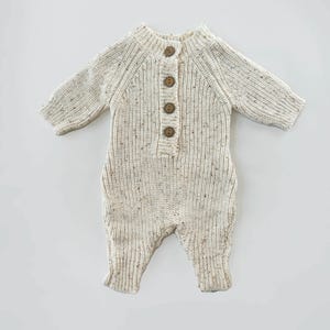 May include: A cream-colored knitted baby onesie with brown button closures. The onesie has a speckled pattern and long sleeves.
