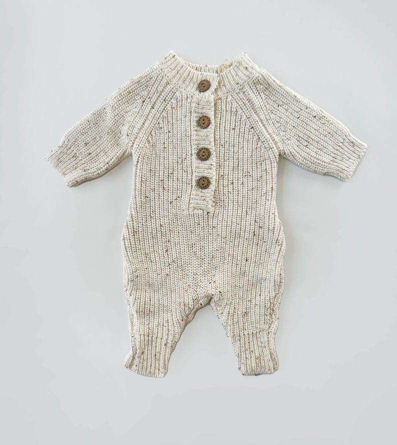 May include: A cream-colored knitted baby onesie with brown button closures. The onesie has a speckled pattern and long sleeves.