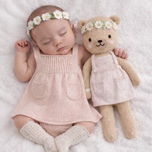 May include: A sleeping baby wearing a light pink knitted dress with two front pockets, and white lace socks. The baby is wearing a floral headband and is next to a matching teddy bear wearing a similar dress and floral headband.