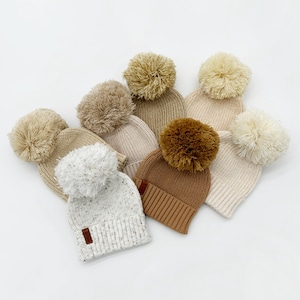 May include: Five knitted hats with pom-poms in various shades of beige and brown. The hats are made of a soft, textured yarn and have a classic beanie style.