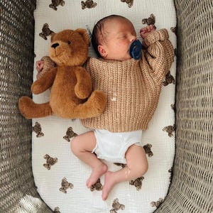 May include: A newborn infant wearing a tan cable-knit sweater and a white diaper, lying on a white muslin sheet patterned with small brown teddy bears. A plush brown teddy bear rests beside the infant, who has a dark blue pacifier in their mouth.