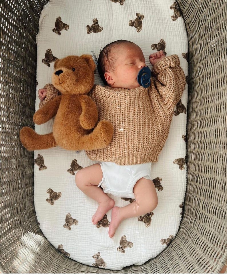 May include: A newborn infant wearing a tan cable-knit sweater and a white diaper, lying on a white muslin sheet patterned with small brown teddy bears. A plush brown teddy bear rests beside the infant, who has a dark blue pacifier in their mouth.