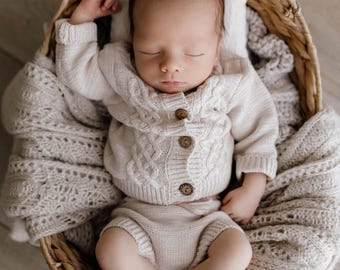 May include: A sleeping baby wearing a cream-colored cable knit sweater and shorts set with wooden buttons, nestled in a woven basket. The baby is lying on a small pillow and covered with a soft blanket. The basket is on a wooden floor.