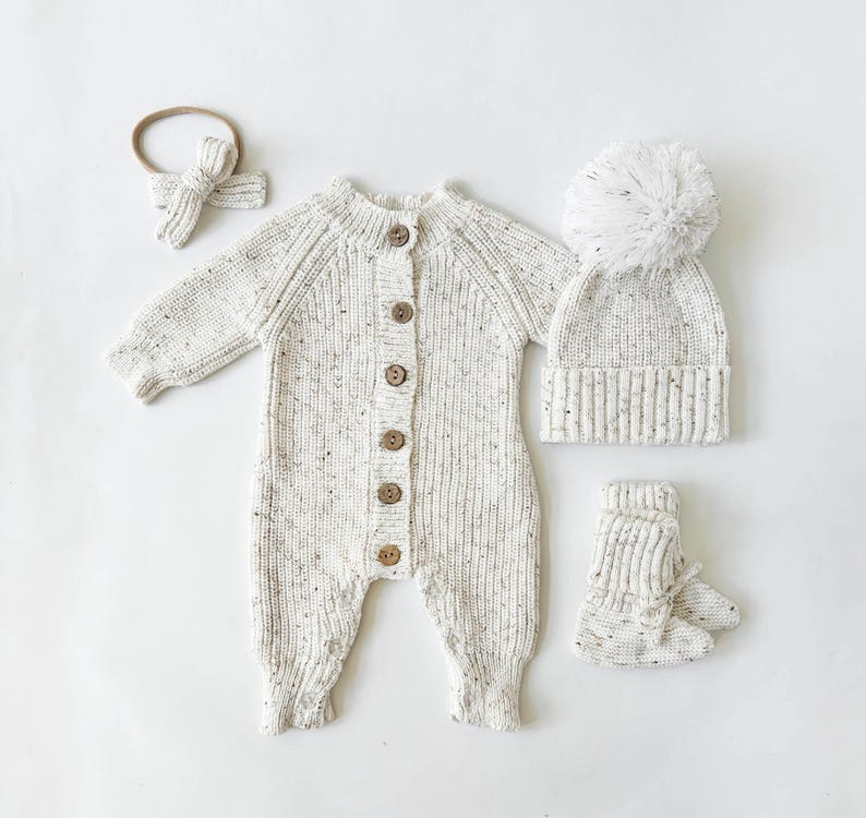 May include: A white knitted baby outfit set with a matching hat, booties, and headband. The outfit features a speckled pattern and button closure.