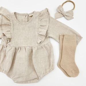 Puede incluir: Un pelele de bebé de lino beige con mangas de volantes, una diadema a juego y un par de calcetines de punto beige.
