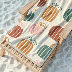 May include: A beach towel with a repeating pattern of colorful pumpkins in shades of orange, pink, green, and brown. The towel is draped over a wooden beach chair, set against a sandy background.