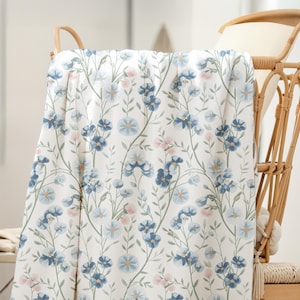 May include: A white blanket with a floral pattern of blue and pink flowers, draped over a wicker chair. The blanket's design includes green stems and leaves. The chair is made of woven wicker.