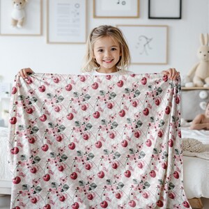 Pode incluir: Um cobertor de beb&ecirc; de tecido branco e rosa com um padr&atilde;o repetido de cerejas vermelhas e flores rosas. O cobertor est&aacute; sendo segurado por uma menina sorridente.