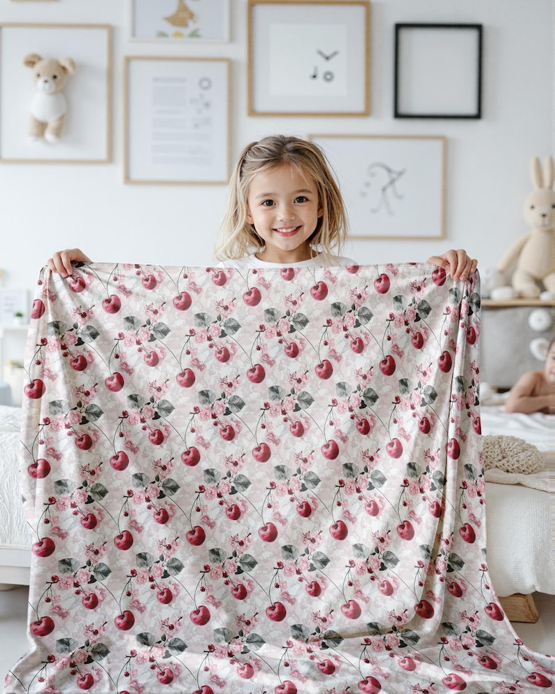 Pode incluir: Um cobertor de beb&ecirc; de tecido branco e rosa com um padr&atilde;o repetido de cerejas vermelhas e flores rosas. O cobertor est&aacute; sendo segurado por uma menina sorridente.