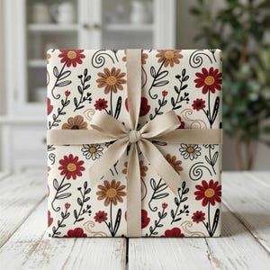 Puede incluir: Una caja de regalo envuelta en papel estampado floral con flores rojas, marrones y blancas, tallos negros y una cinta beige atada con un lazo. La caja está sobre una superficie de madera blanca.